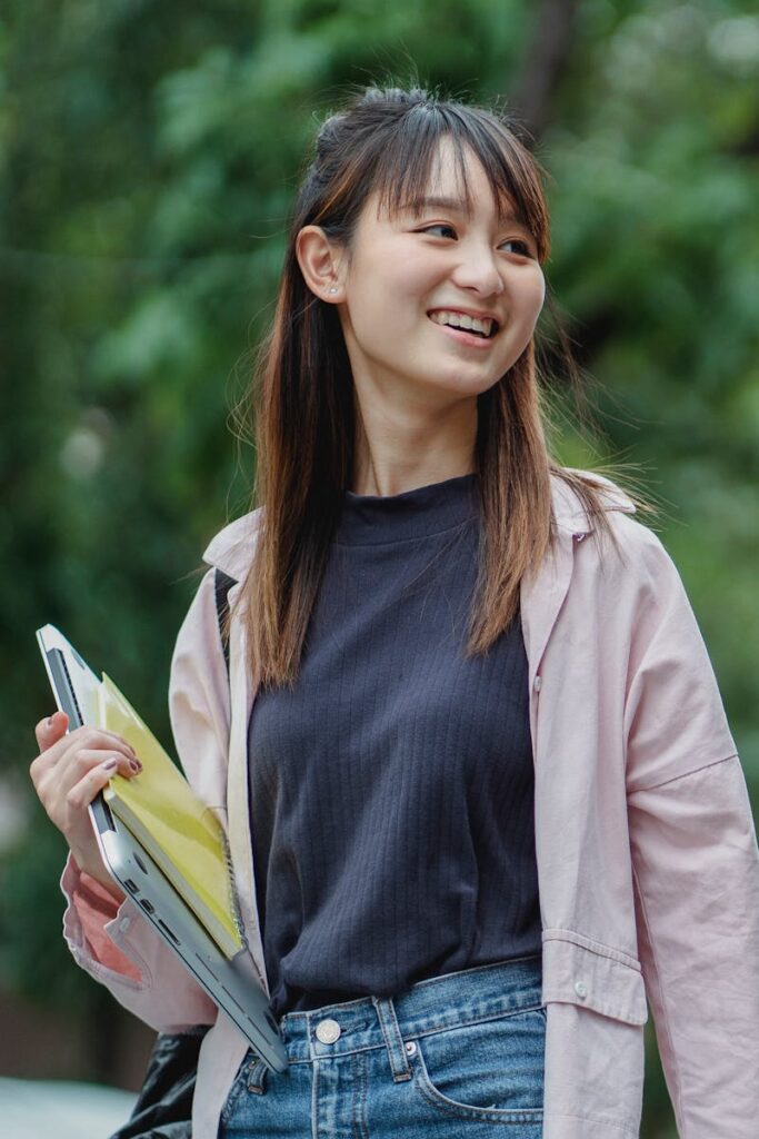 Smiling young Asian woman carrying a laptop and books, enjoying a sunny day outdoors.